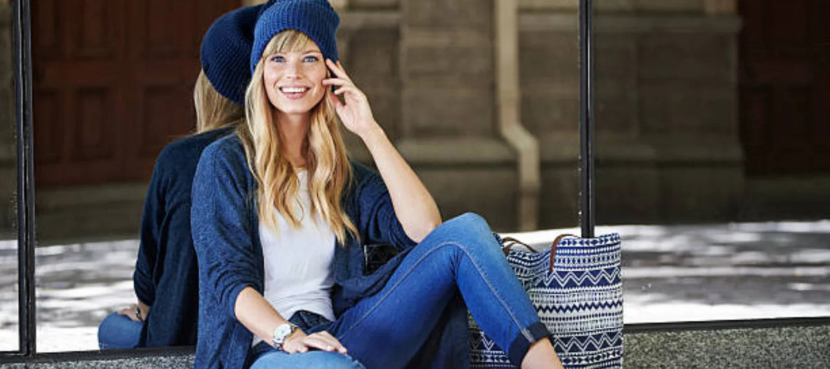 Femme souriante en cardigan bleu, jean et bonnet, assise à l'extérieur avec un sac à motif
