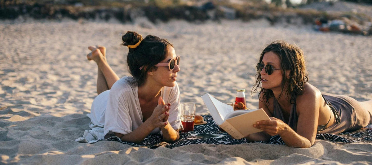 Deux femmes portant des lunettes de soleil tendance, allongées sur une plage, lisant et discutant.