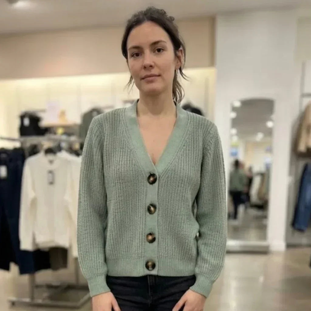 Femme portant un gilet en maille côtelée vert pâle avec boutons marrons dans une boutique.