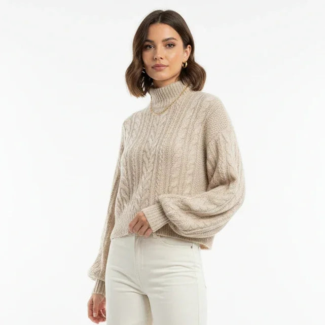 Femme portant un pull en maille torsadée beige à col montant et un pantalon blanc.