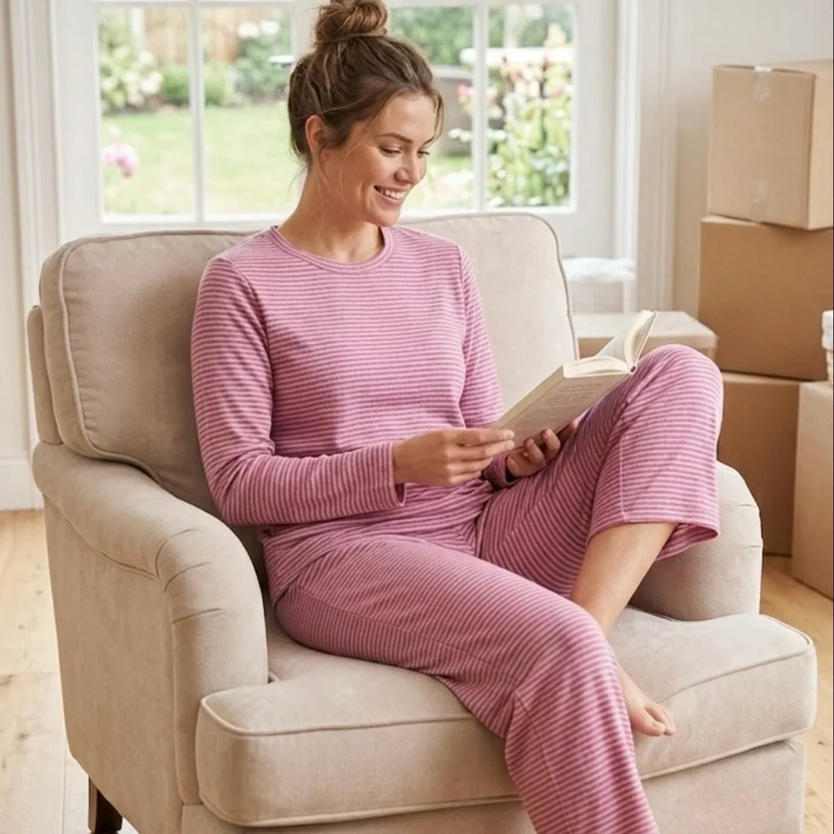 Femme souriante en pyjama rayé rose lisant un livre dans un fauteuil beige
